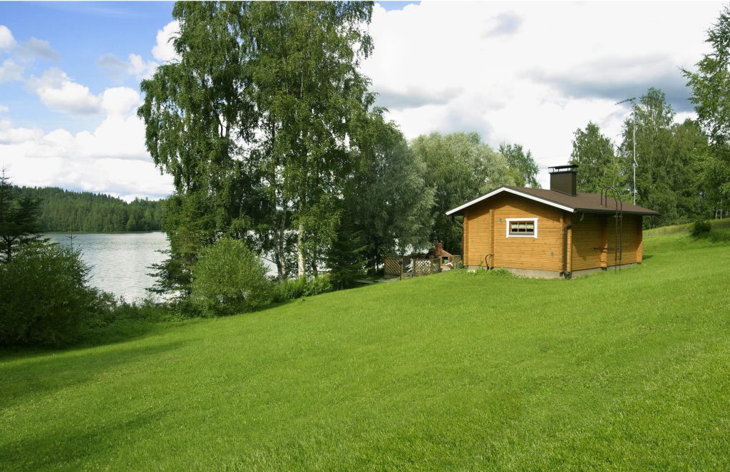 Vihreä siirtonurmialue rantamökin pihalla, taustalla järvi ja metsää. Nurmikko ulottuu rakennukselta alas rantaan, luoden siistin ja luonnollisen ympäristön.