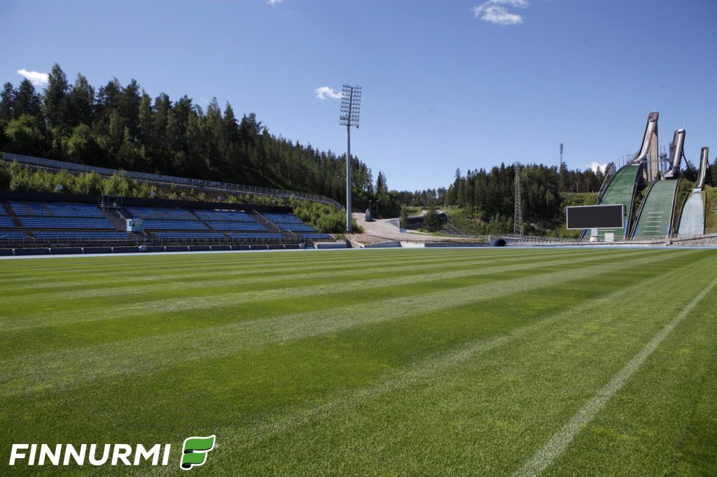 Vihreä, raidallinen siirtonurmikenttä urheilustadionilla, jonka taustalla näkyy katsomo, metsää ja useita hyppyrimäkiä. Kirkas kesäpäivä ja sininen taivas korostavat kentän värejä. Vasemmassa alakulmassa Finnurmi-logo.