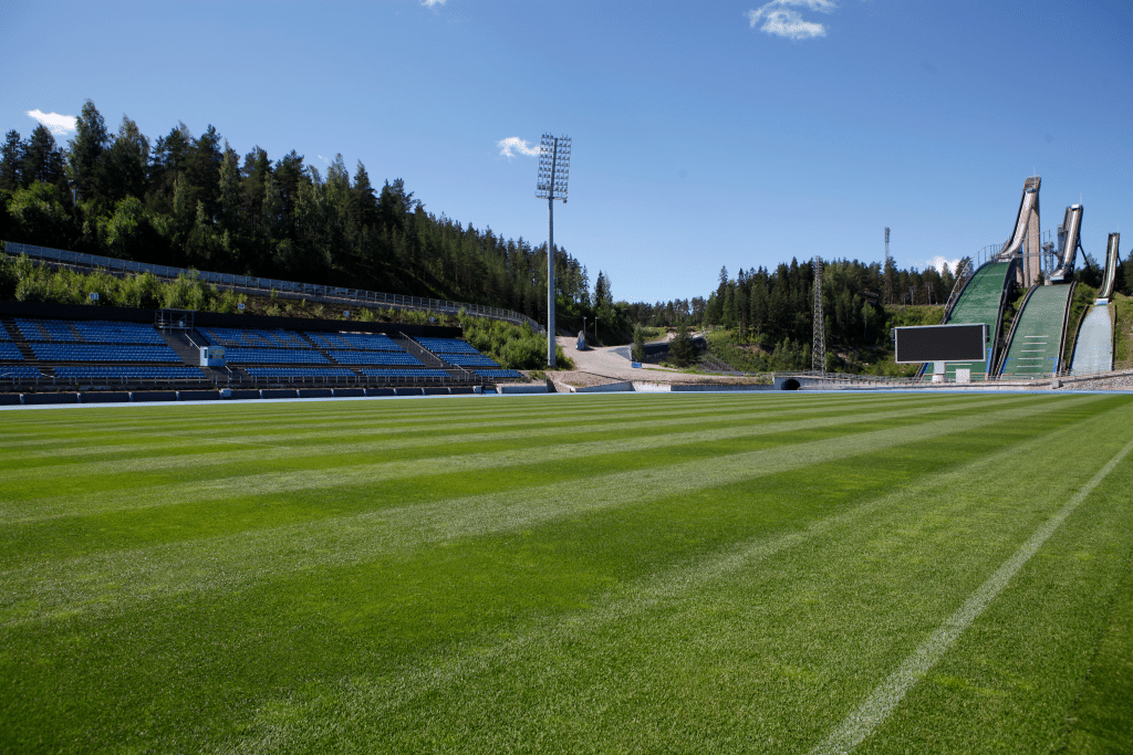 Lahden mäkimonttu kesäasussaan, jossa stadionin kenttä on päällystetty tiheällä ja tasaisella siirtonurmella. Taustalla näkyvät hyppyrimäet ja katsomot.