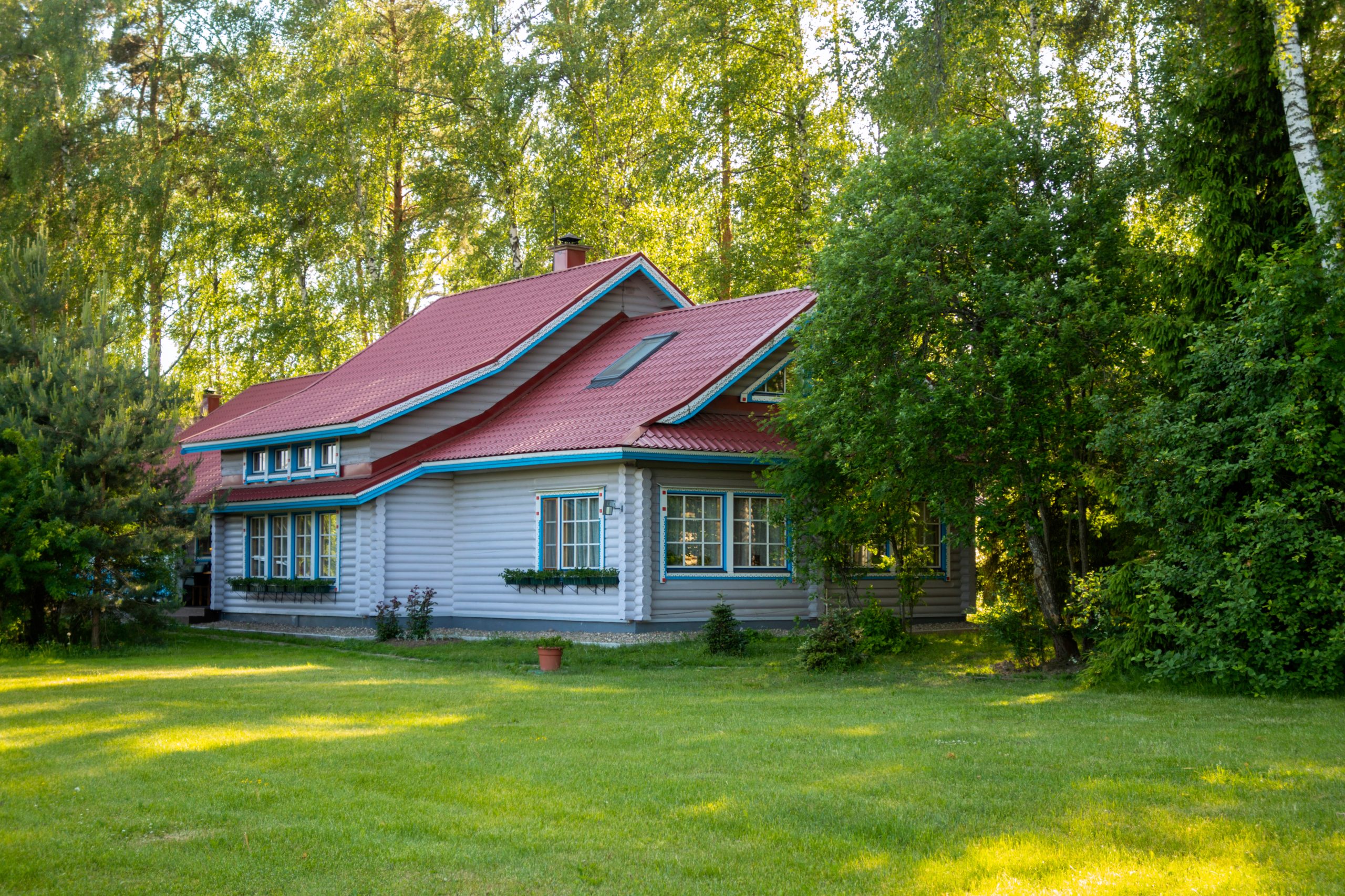 Vihreän nurmikon ympäröimä vaalea omakotitalo punaisella katolla metsäisessä maisemassa.