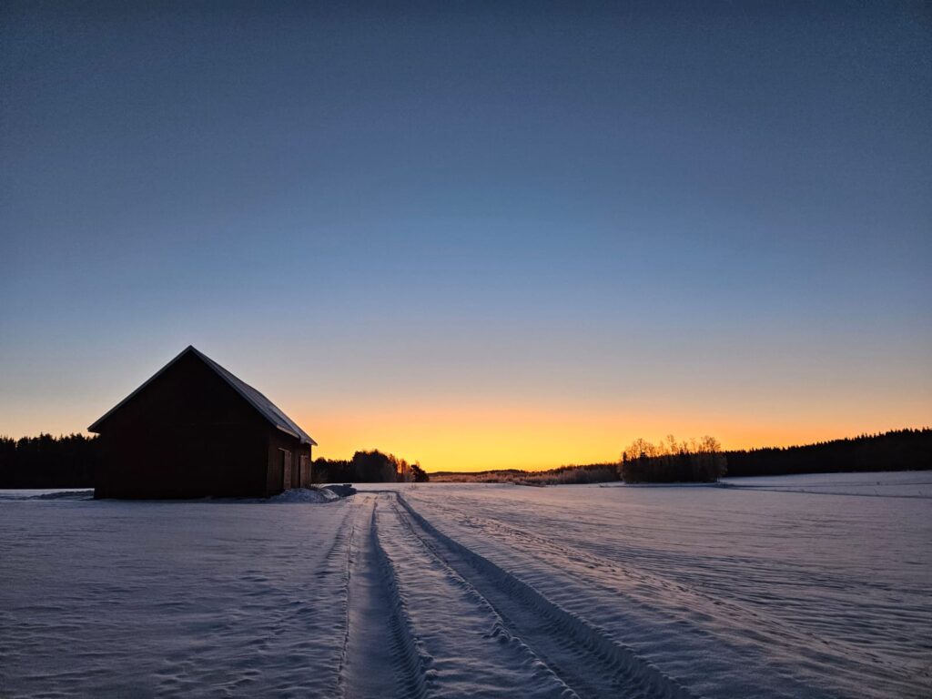 Lato ja luminen pelto auringonlaskussa – talvinen ilta suomalaisessa maalaismaisemassa.
